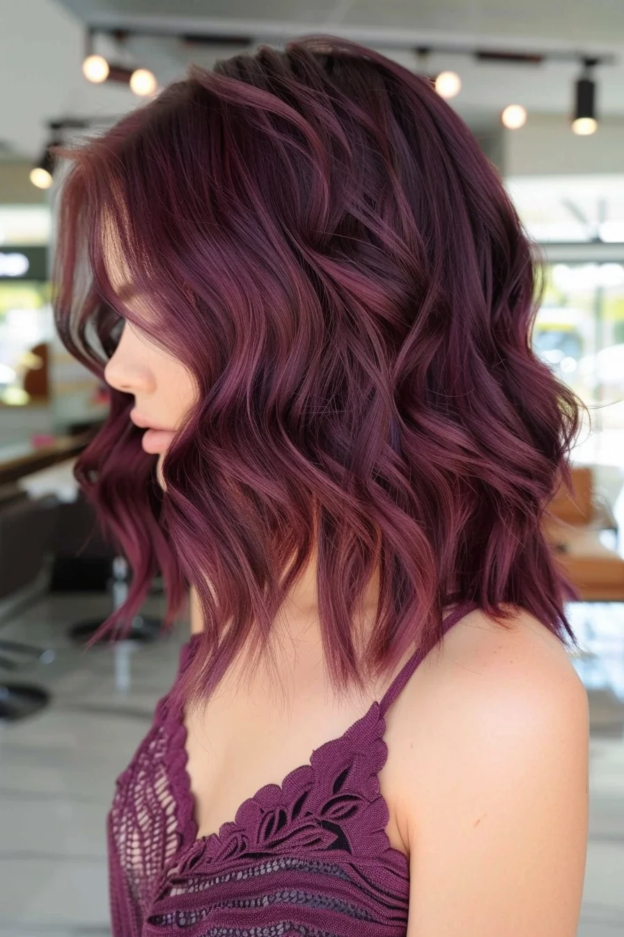 A striking shoulder-length cut features deep wine tones that transition into vibrant burgundy highlights. Waves styled with purposeful texture show off the color's remarkable dimension, while shorter layers around the crown create volume and movement. Darker roots blend flawlessly into brighter mid-lengths and ends, creating a rich, multi-dimensional effect.